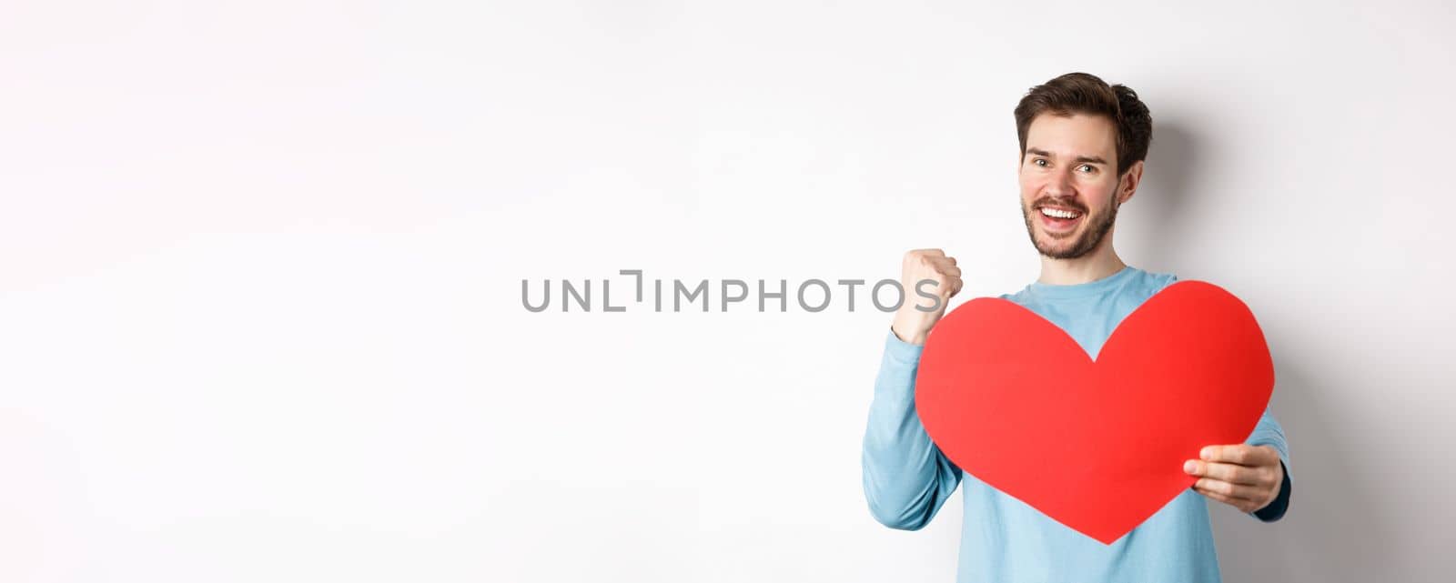 Valentines day. Happy boyfriend triumphing, saying yes and showing valentine red heart, smiling as winning girls love, standing over white background.