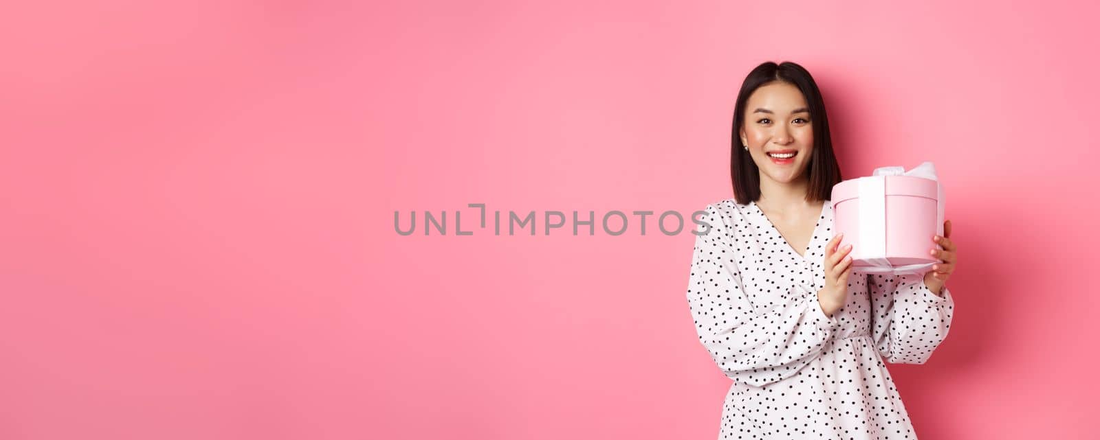 Romantic asian woman in cute dress holding box with gift, smiling happy at camera, standing with present over pink background by Benzoix