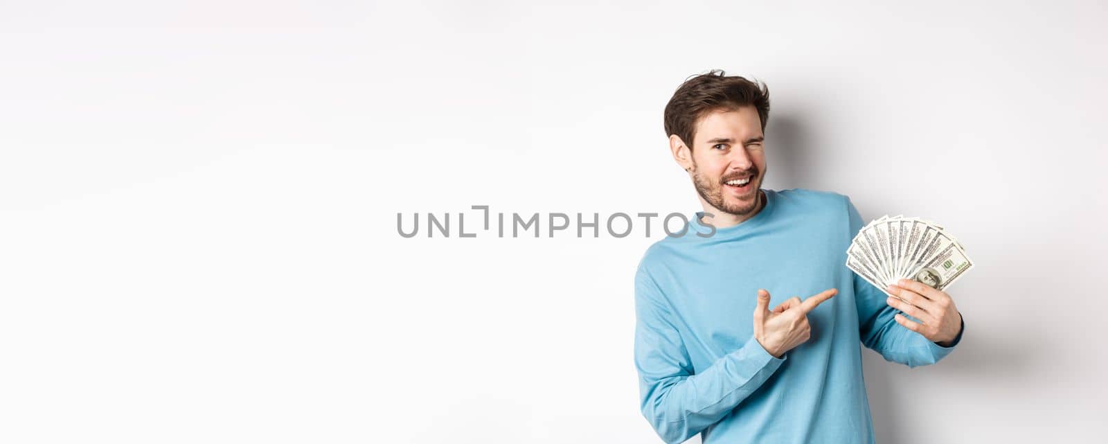 Handsome rich guy winking at camera, showing income, pointing finger at earned money and smiling, standing over white background.