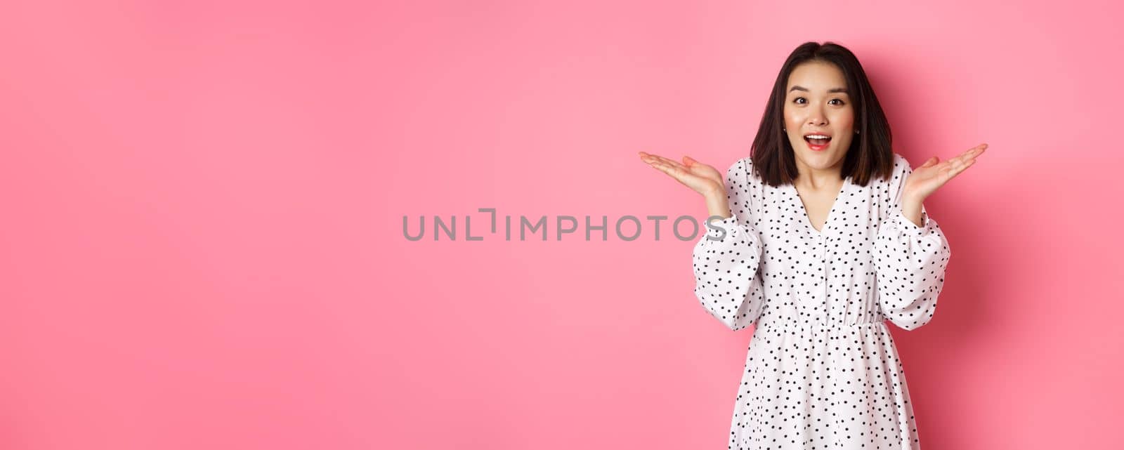 Image of surprised korean girl in dress, female model staring at camera and gasping amazd, standing over pink background.
