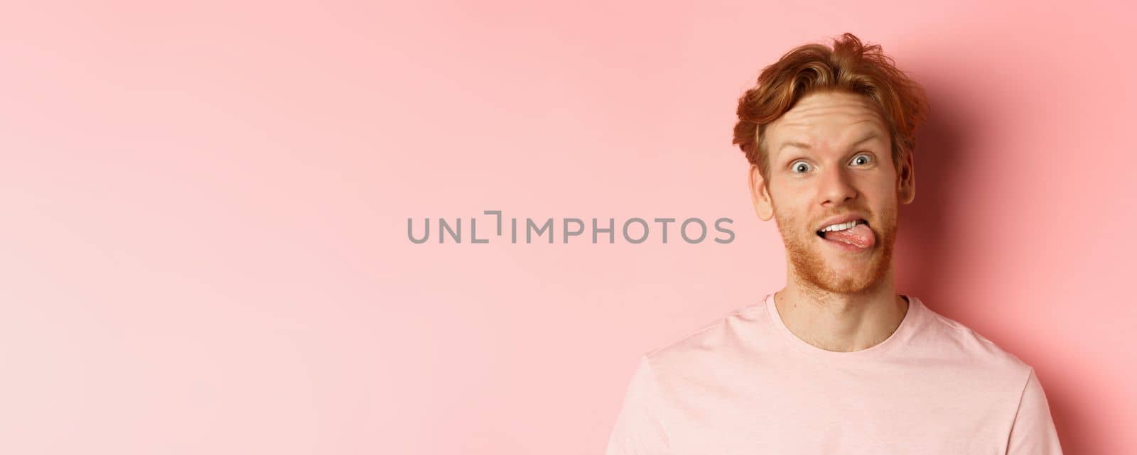 Headshot of funny redhead guy showing tongue, making silly faces at camera, standing joyful against pink background.