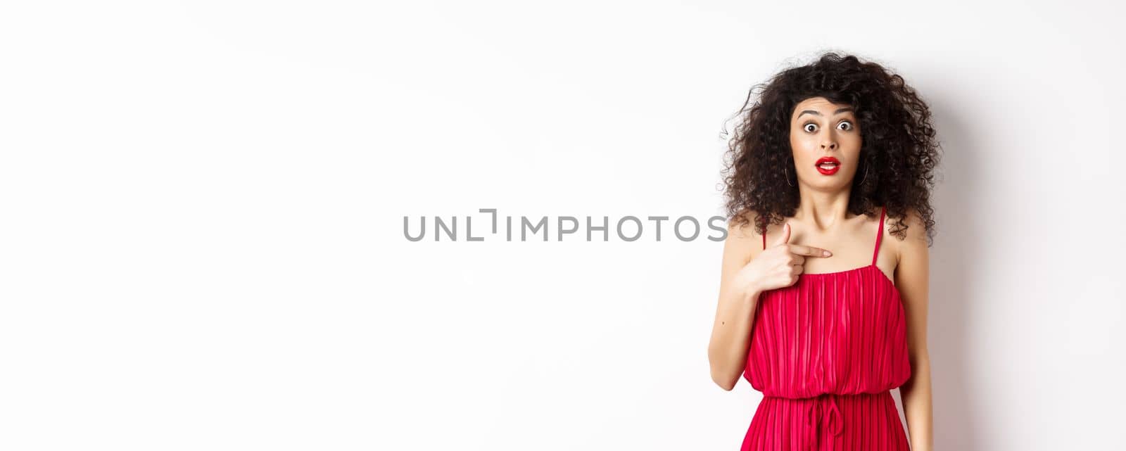 Surprised young woman in red dress pointing at herself, being confused and shocked, standing on white background by Benzoix