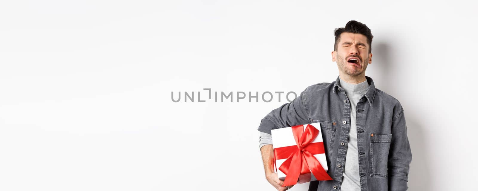 Valentines day heartbreak. Single and heartbroken guy crying lonely, holding big gift box, being rejected by lover, sobbing and feeling alone, standing on white background by Benzoix