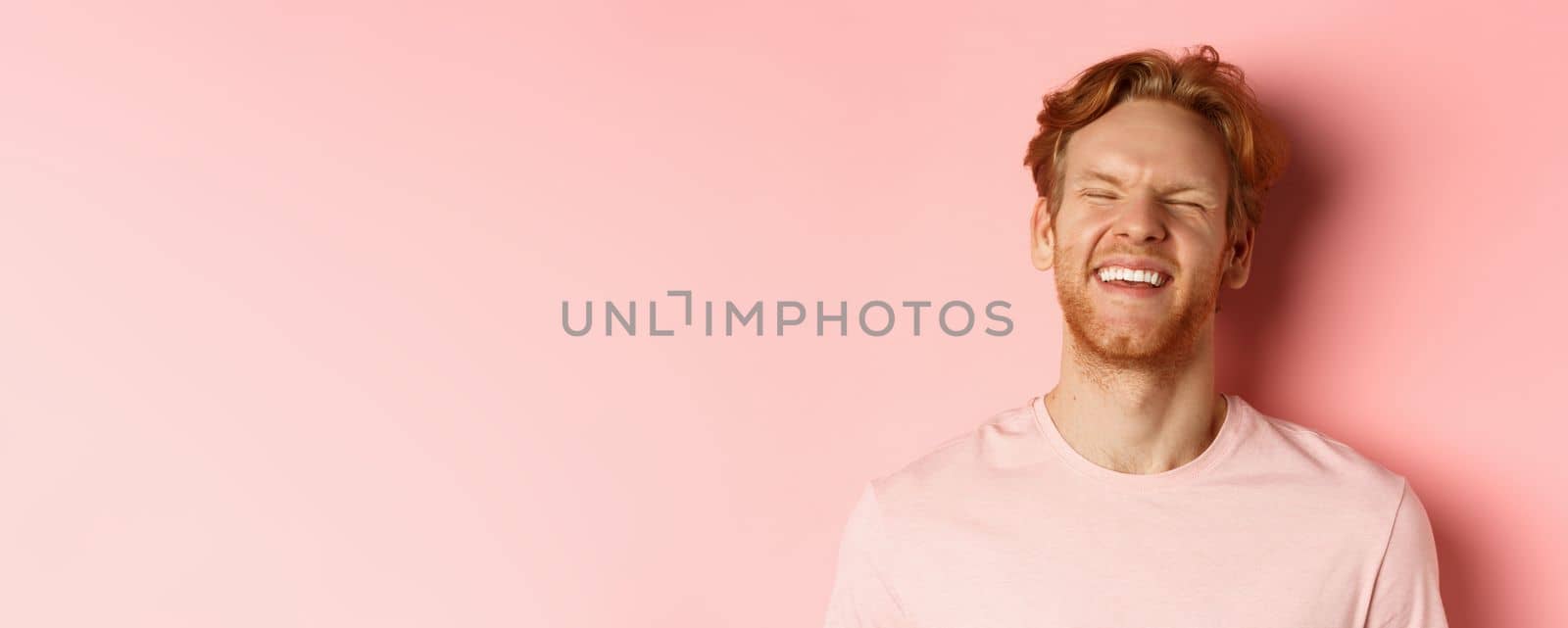 Close up of carefree man with red hair and beard, laughing and feeling happy, standing over pink background, having fun.