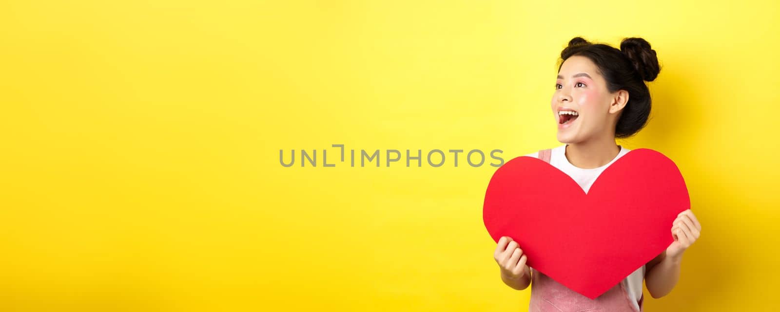 Happy valentines and love concept. Cheerful japanese girl looking left, showing big red heart and smiling happy at lover, standing on yellow background.