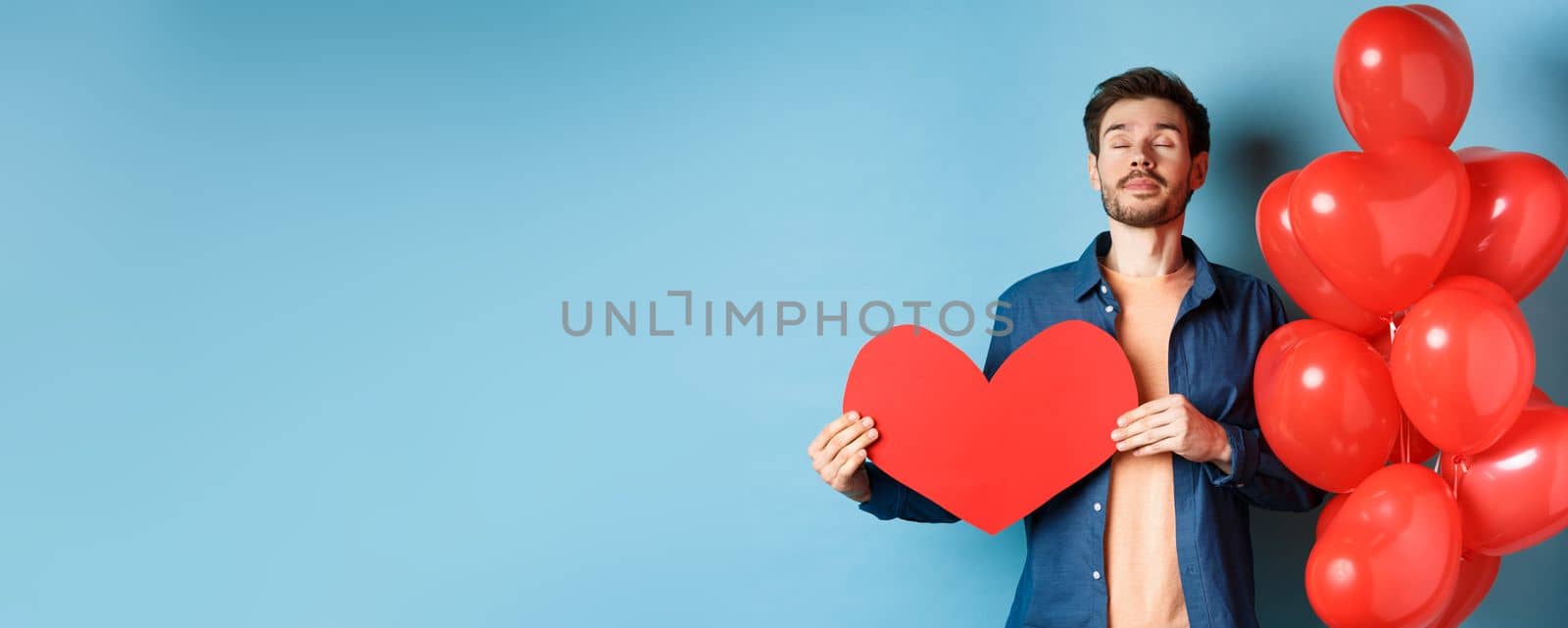 Valentines day concept. Man dreaming of true love, holding red heart cutout and standing near romantic balloons, blue background.