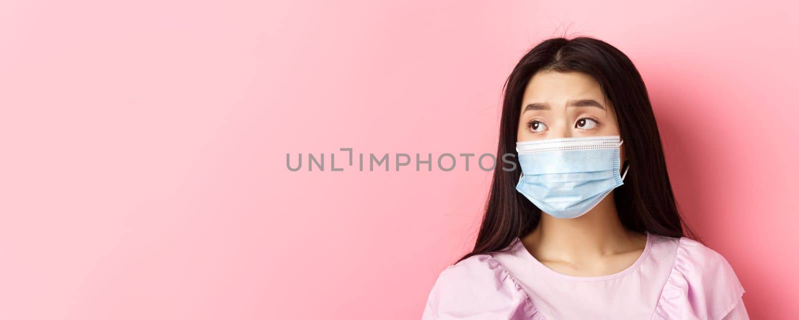 Covid-19, pandemic and quarantine concept. Close-up of sad asian girl in medical mask feeling lonely during coronavirus, standing against pink background by Benzoix