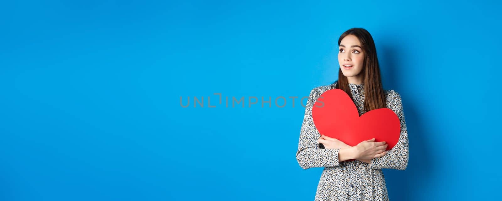 Valentines day. Hopeful girl dreaming off soulmate, looking aside at empty space with excitement, hugging big red heart cutout, standing on blue background.