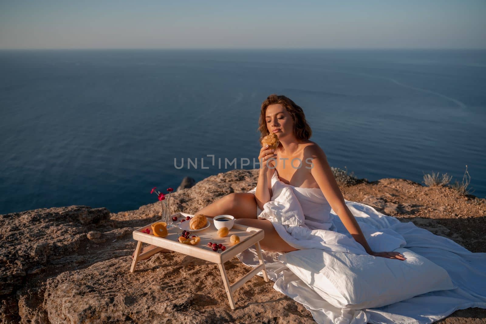 Woman covered with a blanket of bed relaxing and watching the seascape at sunrise. She holds a cup of coffee in her hand in front of her is a table with fruits and croissants. Wanderlust and freedom concept scene