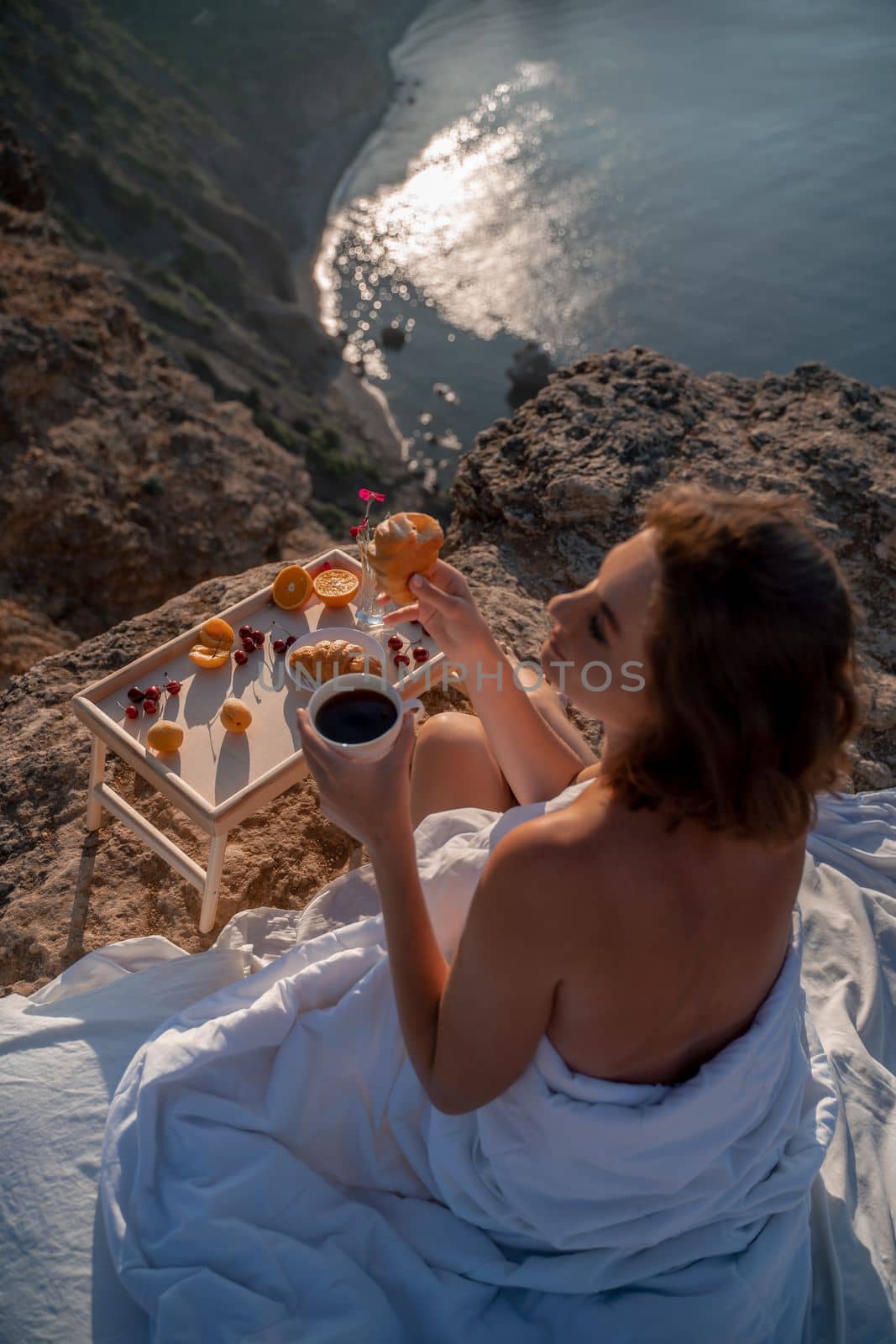 Woman covered with a blanket of bed relaxing and watching the seascape at sunrise. She holds a cup of coffee in her hand in front of her is a table with fruits and croissants. Wanderlust and freedom concept scene. by Matiunina