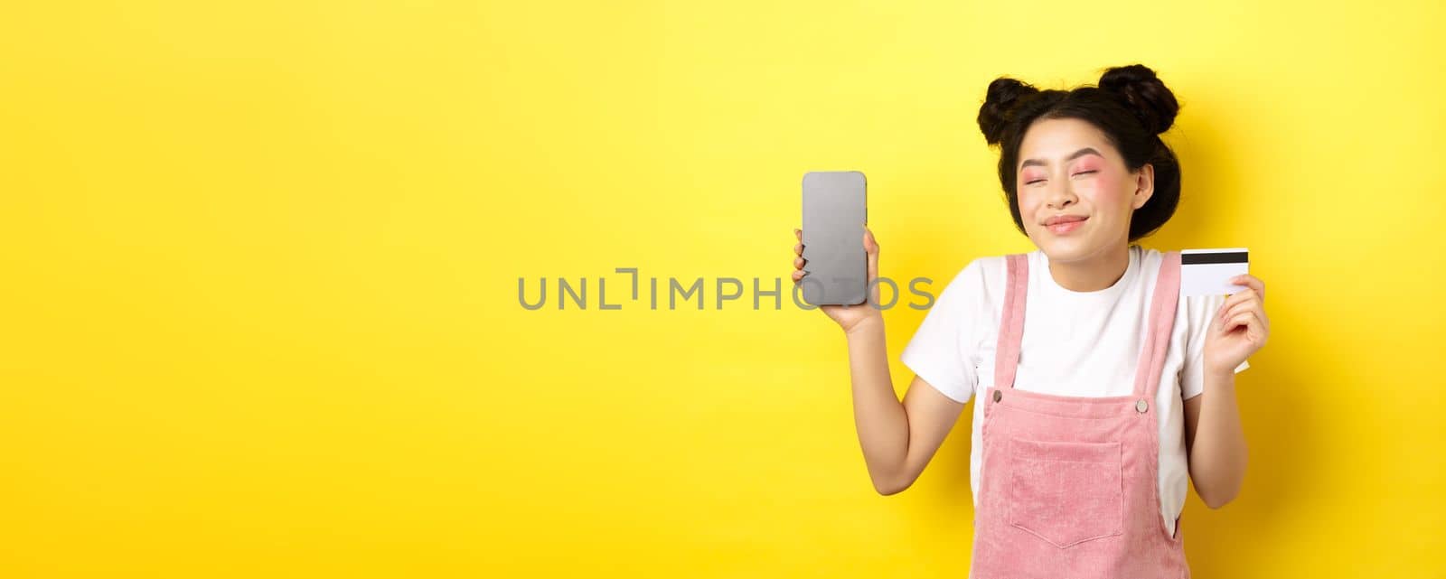 Online shopping concept. Happy asian girl showing empty smartphone screen and credit card, paying contactless, standing on yellow background.