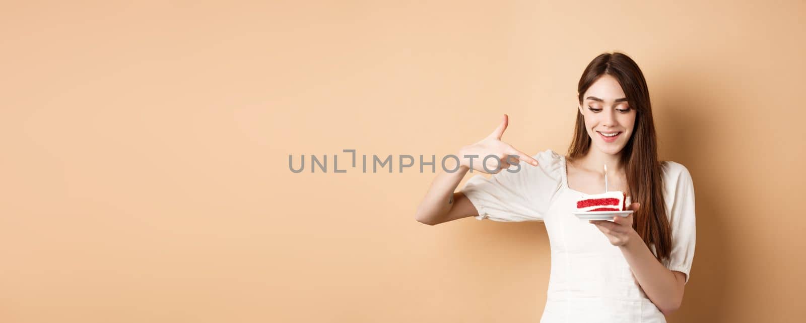 Cute girl pointing at birthday cake with candle and smiling excited, making bday wish, standing on beige background.