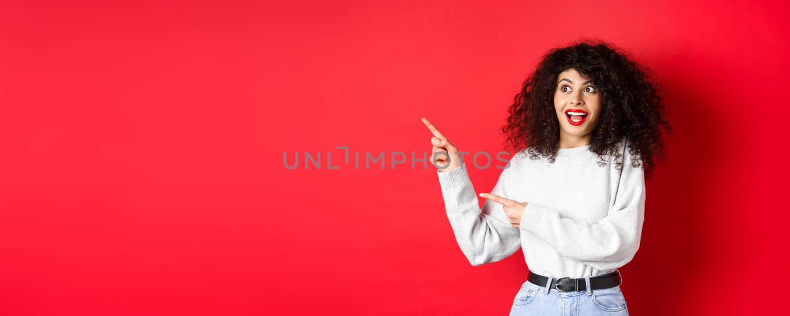 Excited pretty girl with curly hair and red lips, looking and pointing fingers left with amazed face, showing banner, standing on studio background by Benzoix