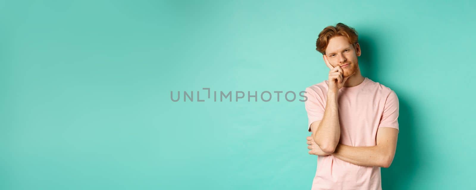 Bored handsome guy with red hair leaning on hand, looking tired and unamused at camera, standing in t-shirt against turquoise background by Benzoix