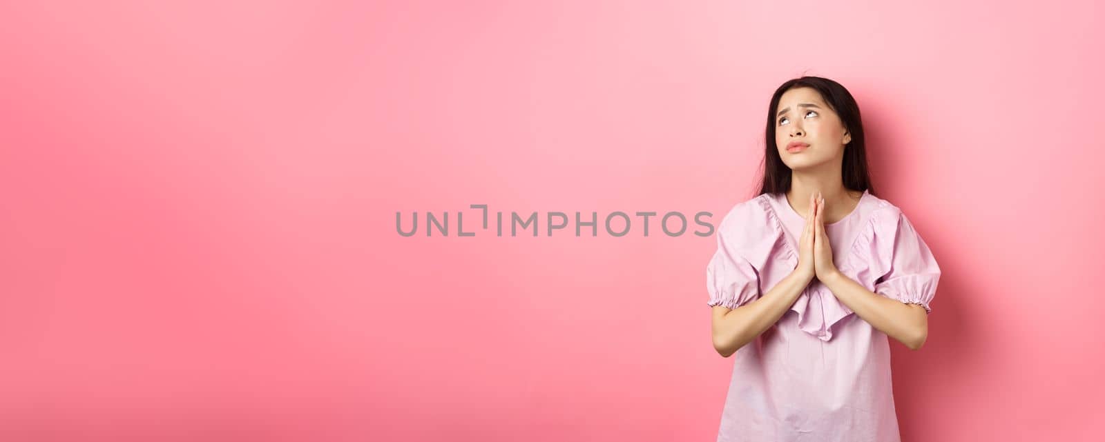 Hopeful asian girl praying god, looking up to plead or pray, making wish, supplicating with dramatic face, standing against pink background by Benzoix