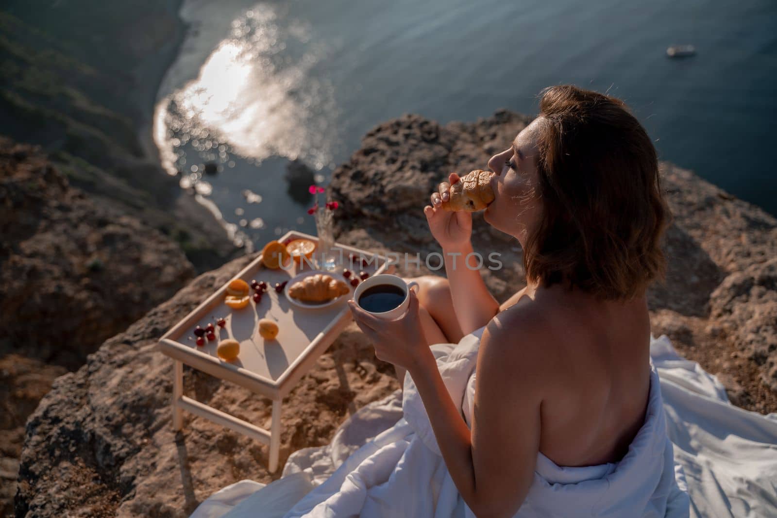 Woman covered with a blanket of bed relaxing and watching the seascape at sunrise. She holds a cup of coffee in her hand in front of her is a table with fruits and croissants. Wanderlust and freedom concept scene. by Matiunina