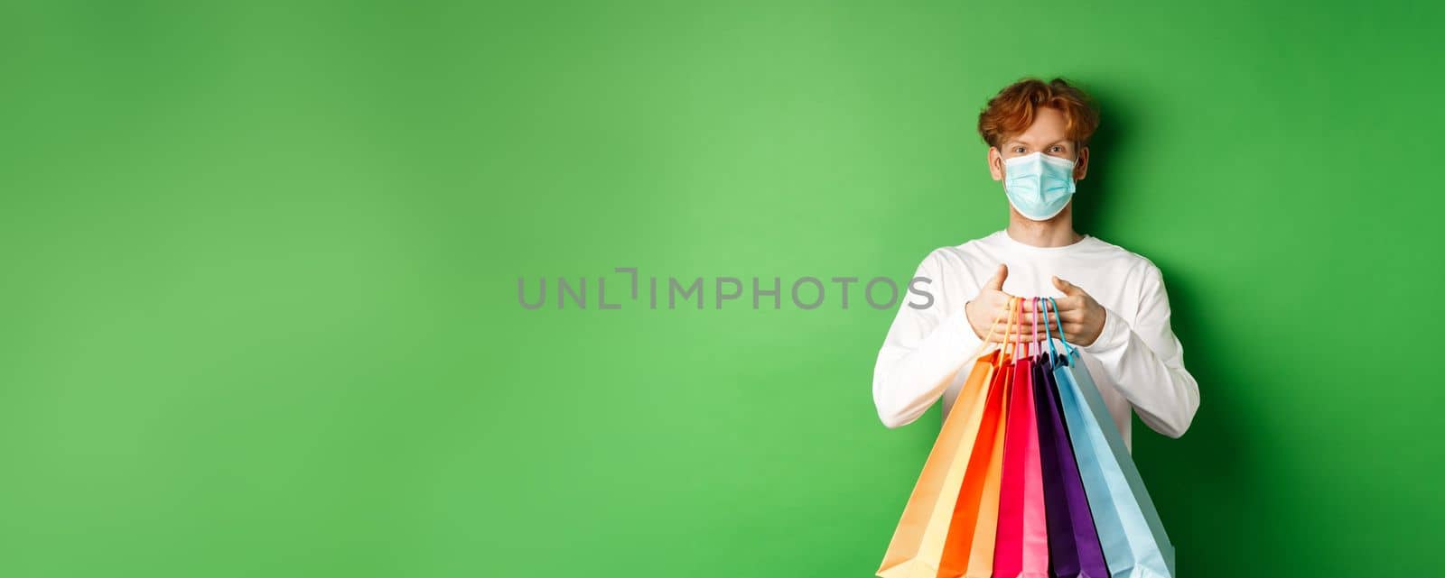 Pandemic and lifestyle concept. Cheerful redhead man going shopping in store, wearing medical mask and holding bags, standing over green background.