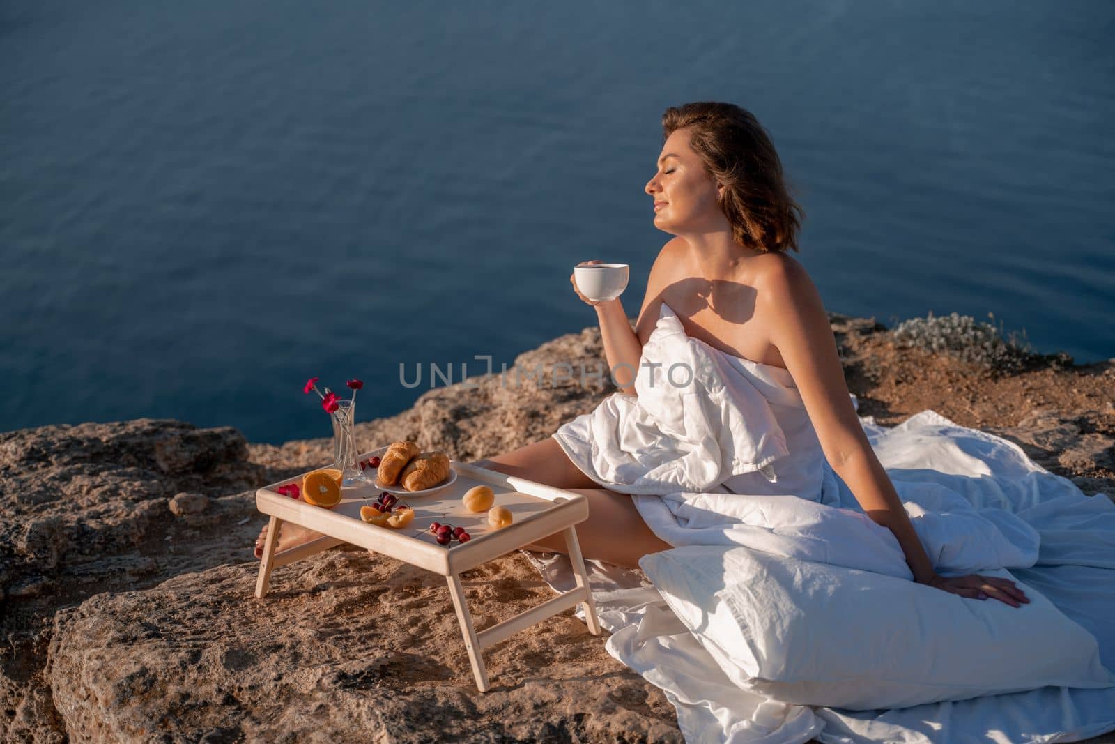 Woman covered with a blanket of bed relaxing and watching the seascape at sunrise. She holds a cup of coffee in her hand in front of her is a table with fruits and croissants. Wanderlust and freedom concept scene. by Matiunina