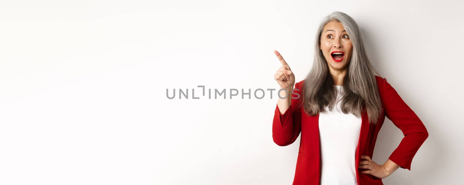 Surprised senior asian lady pointing upper left corner, checking out promo offer and smiling amazed, standing in red blazer over white background by Benzoix