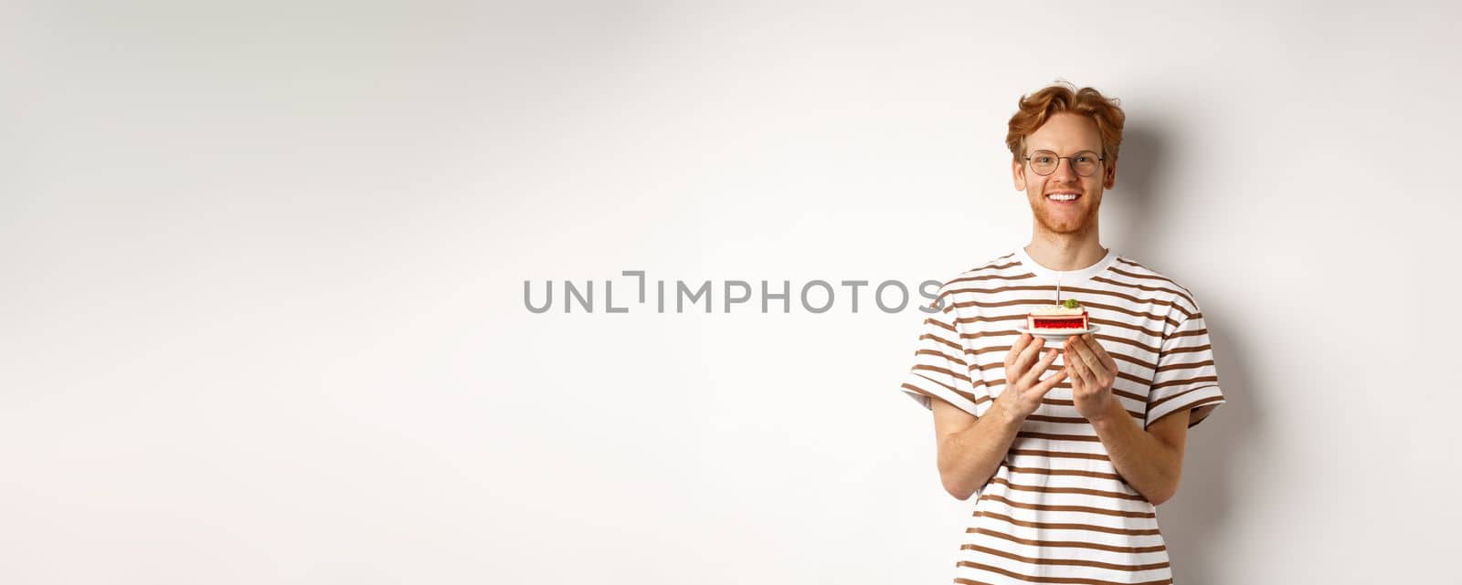 Holidays and celebration concept. Handsome redhead man in glasses holding birthday cake with candle, smiling and looking happy at camera by Benzoix