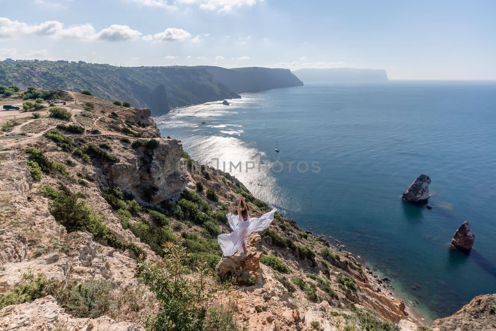 A beautiful young woman in a white light dress with long legs stands on the edge of a cliff above the sea waving a white long dress, against the background of the blue sky and the sea. by Matiunina