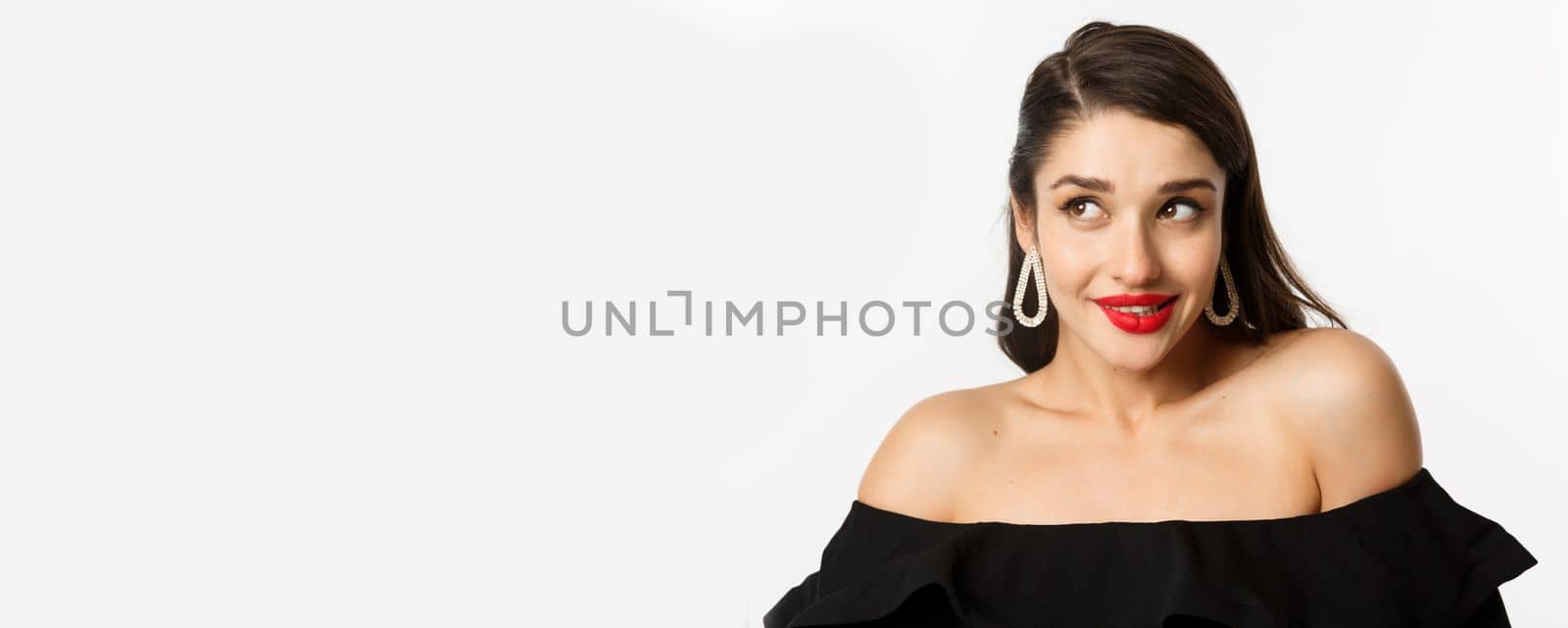 Fashion and beauty concept. Close-up of beautiful woman with red lips, makeup and black dress, showing her earrings and looking pleased, white background.