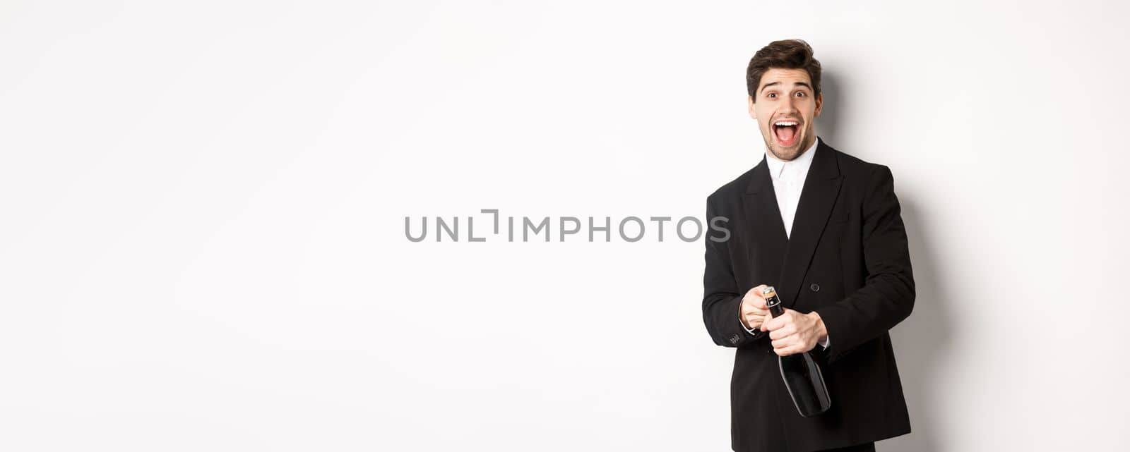Image of attractive man in black suit having a party, celebrating new year and opening bottle of champagne, standing happy against white background by Benzoix