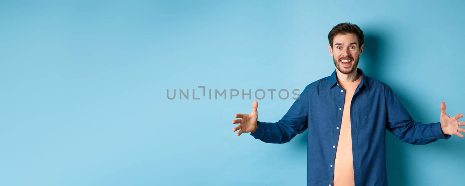 Happy smiling man spread hands sideways and showing big size object, talking about large object, standing cheerful on blue background by Benzoix