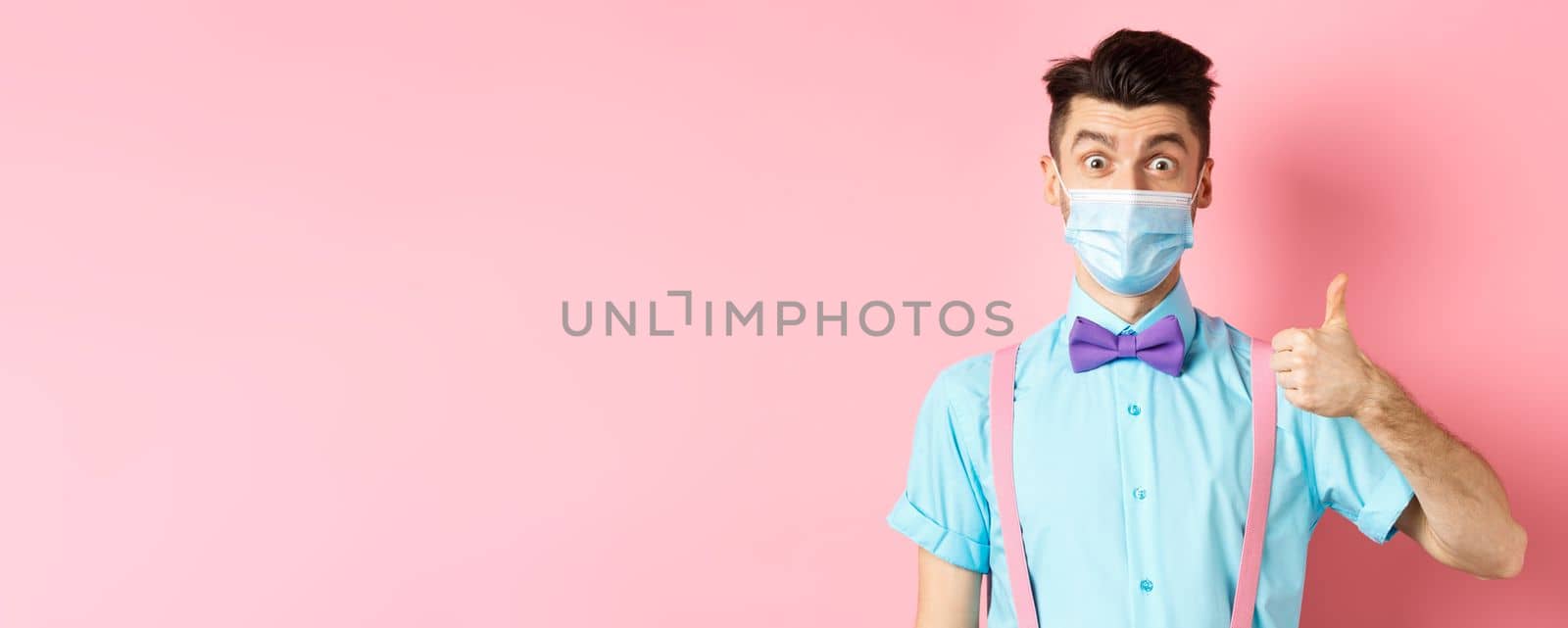 Coronavirus, healthcare and quarantine concept. Impressed young man in medical mask showing thumb up, looking satisfied, recommending good product, pink background.