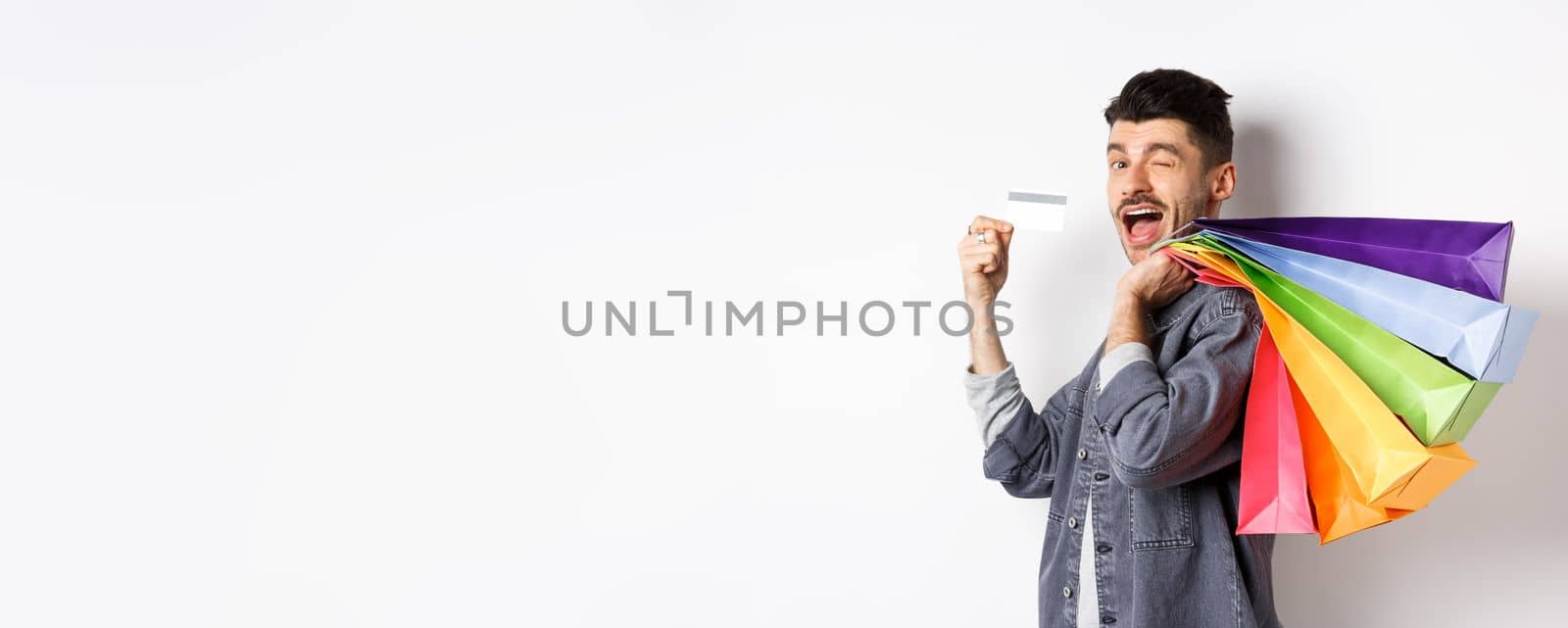 Happy shopper holding shopping bags over shoulder and showing plastic credit card, paying contactless, standing on white background.