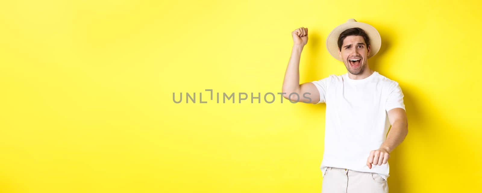 Concept of tourism and summer. Young man traveller showing rodeo gesture, standing in straw hat and white clothes, standing over yellow background.