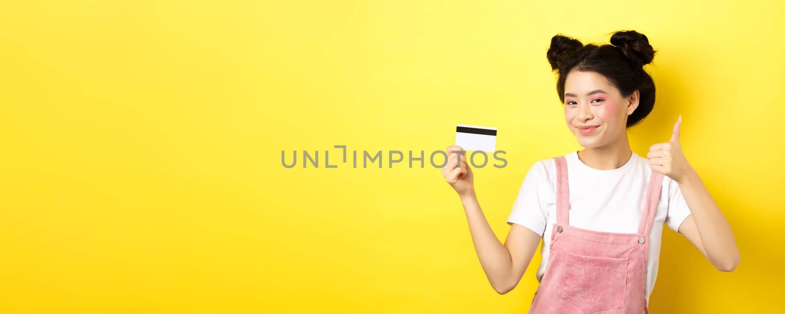 Shopping. Stylish asian glam girl showing thumb up and plastic credit card, standing on yellow background by Benzoix