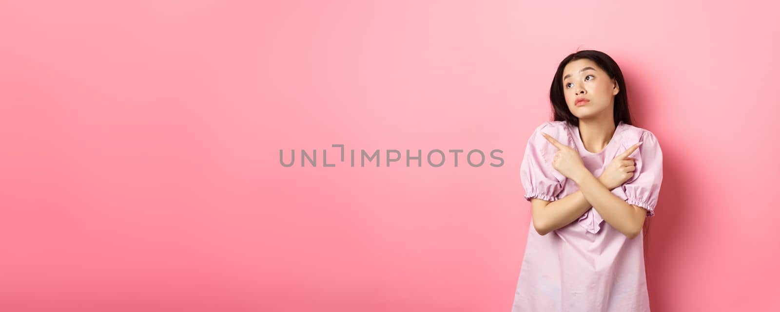 Indecisive asian woman cant decide, pointing fingers sideways and look pensive aside, making choice, standing confused on pink background by Benzoix