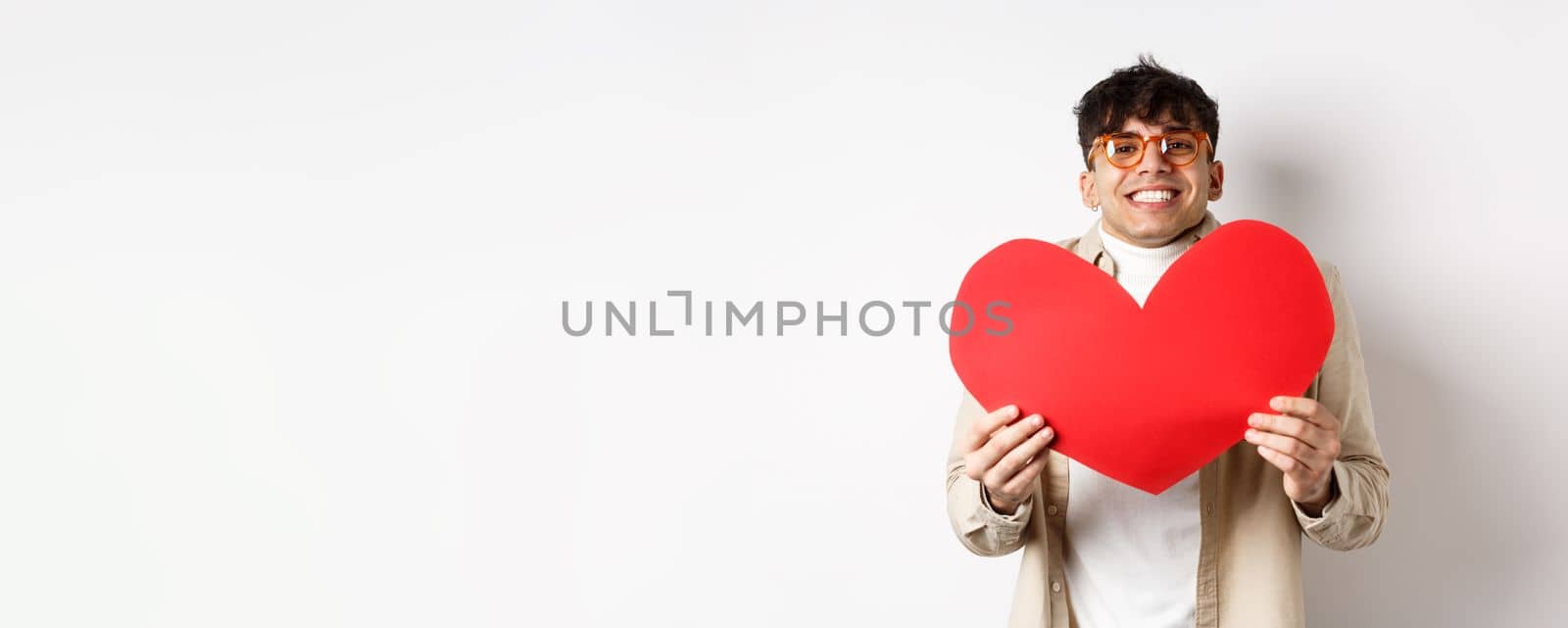 Cheerful young man smiling and looking thrilled at camera, showing big red heart cutout on Valentines day, making romantic gift to lover, white background.