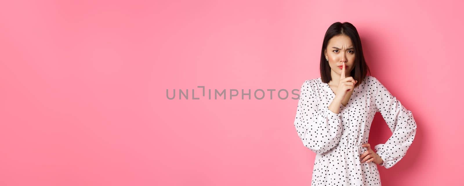 Angry asian female model shushing, frowning and hushing, showing taboo gesture, standing displeased against pink background, demand silence by Benzoix