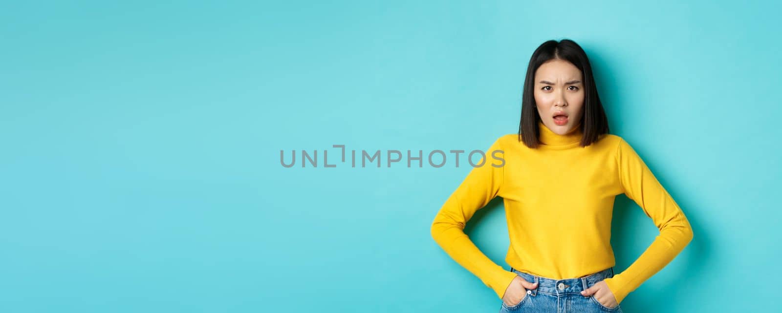 Portrait of disappointed korean woman in yellow sweater, frowning and looking upset, standing over blue background.