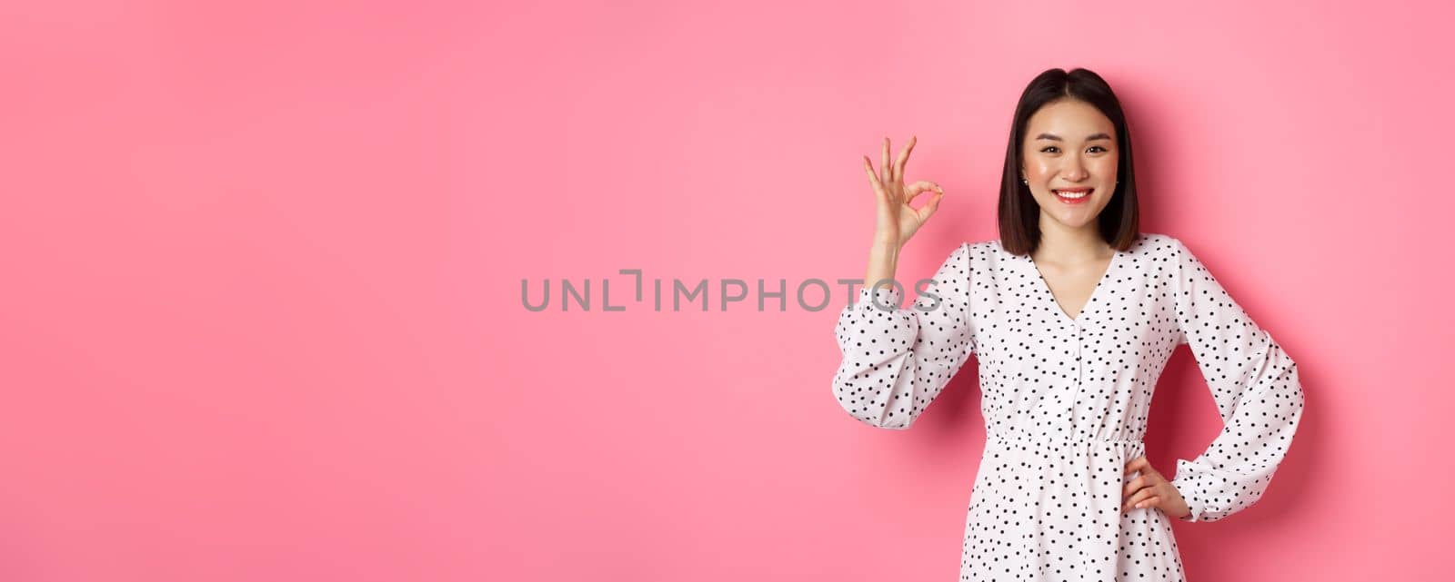 Pretty young asian woman in dress showing okay sign, praising and showing approval, looking satisfied, standing against pink background.