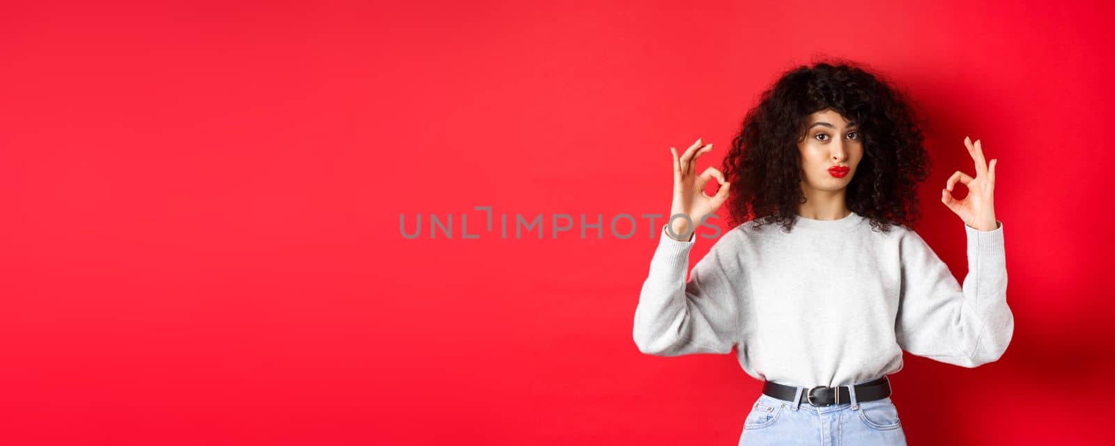 Not bad. Impressed curly woman nod in approval, pucker lips and showing okay signs, like good thing, standing satisfied on red background.