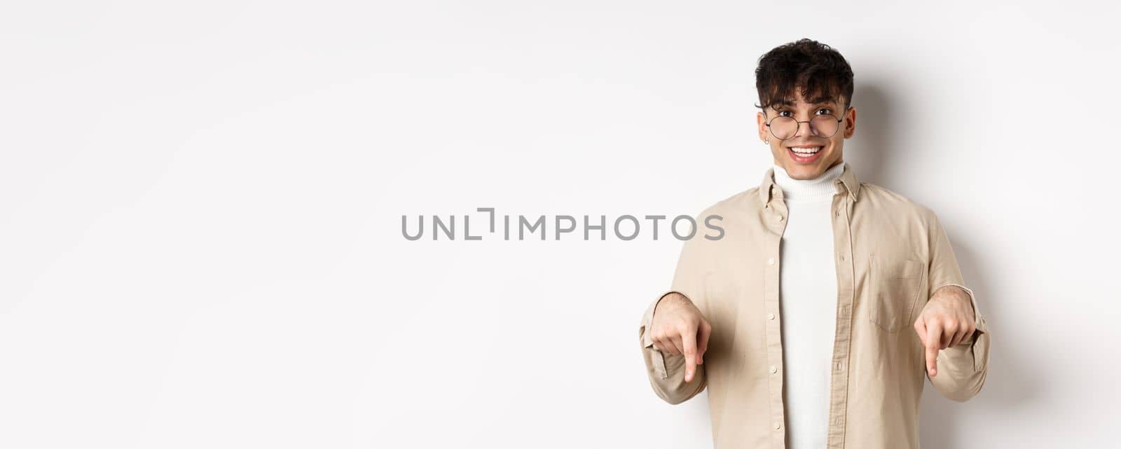 Portrait of happy young guy in glasses showing cool promo, pointing fingers down and smiling excited, standing on white background.