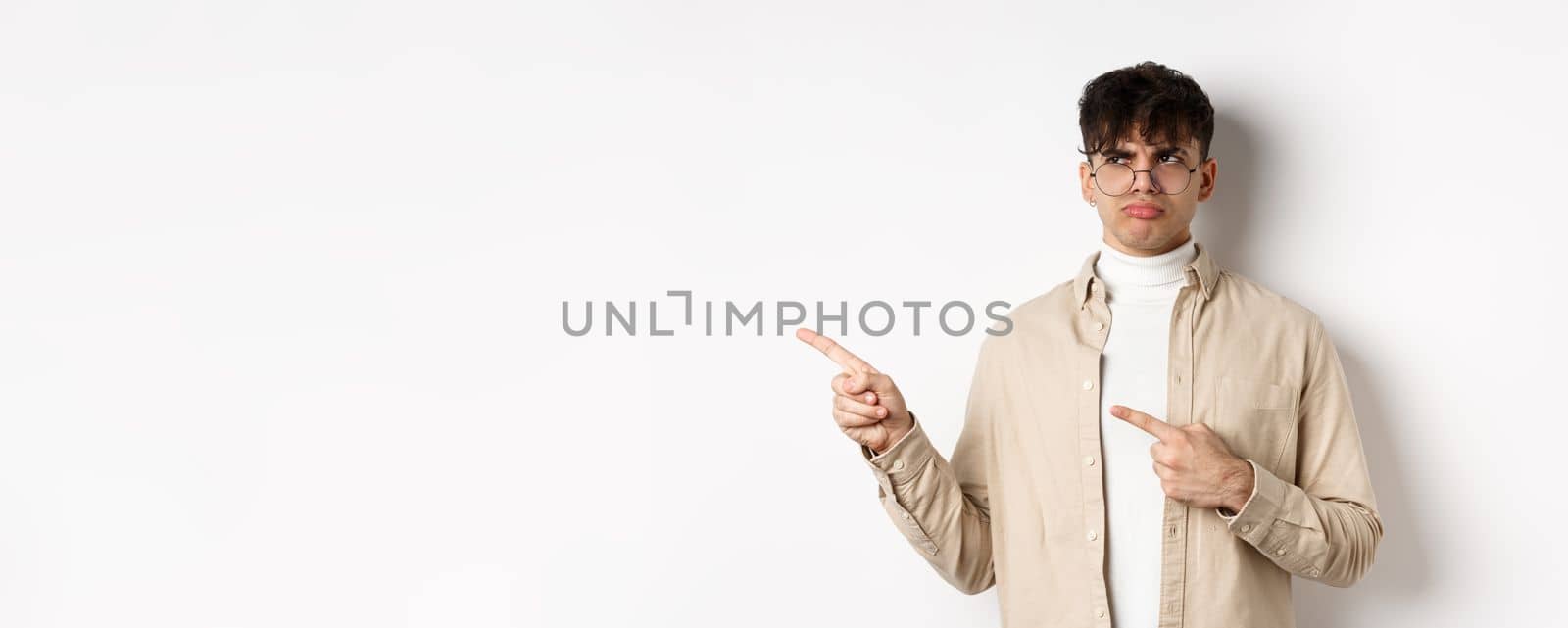 Disappointed guy in glasses looking and pointing left, sulking displeased, complaining on bad offer, standing upset on white background.