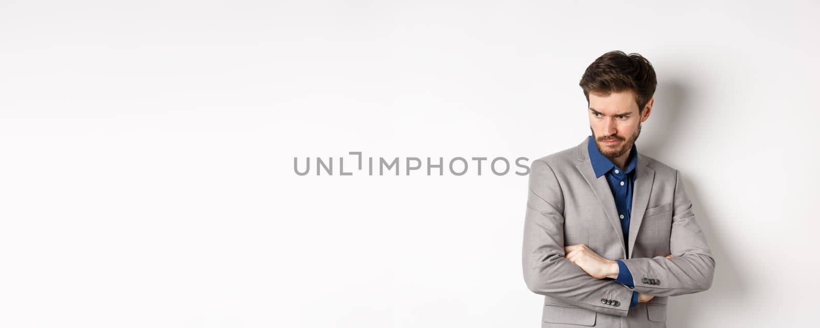 Moody guy in suit looking aside with unfair face, cross arms on chest and frowning angry, standing upset against white background.