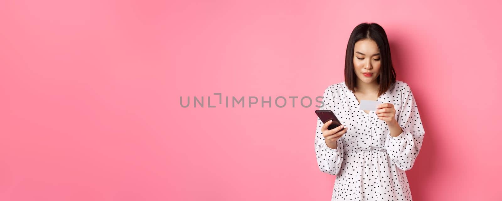 Beautiful asian woman enter credit card info in mobile phone app, shopping online, paying for order on smartphone, standing over pink background.