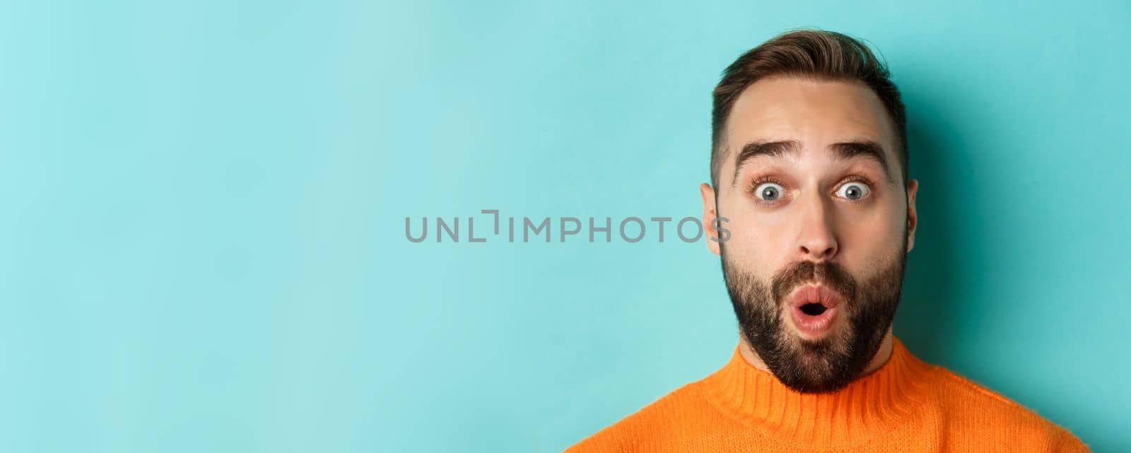 Headshot of handsome caucasian man with beard standing in orange sweater against turquoise background, saying wow and staring surprised at camera.