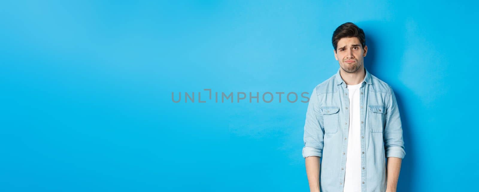 Man looking with doubt at camera, frowning and sulking, feeling uneasy about something, standing against blue background.