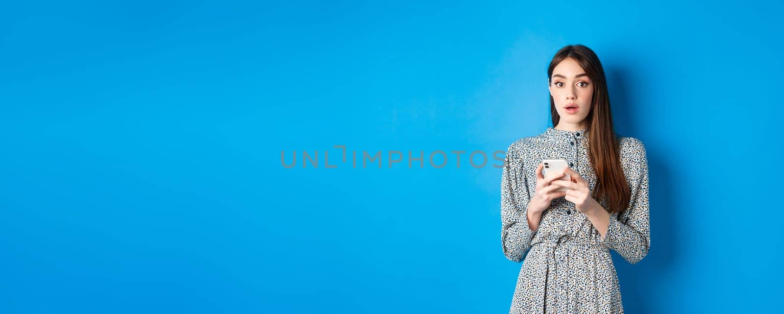 Woman look surprised after using mobile phone, standing on blue background.