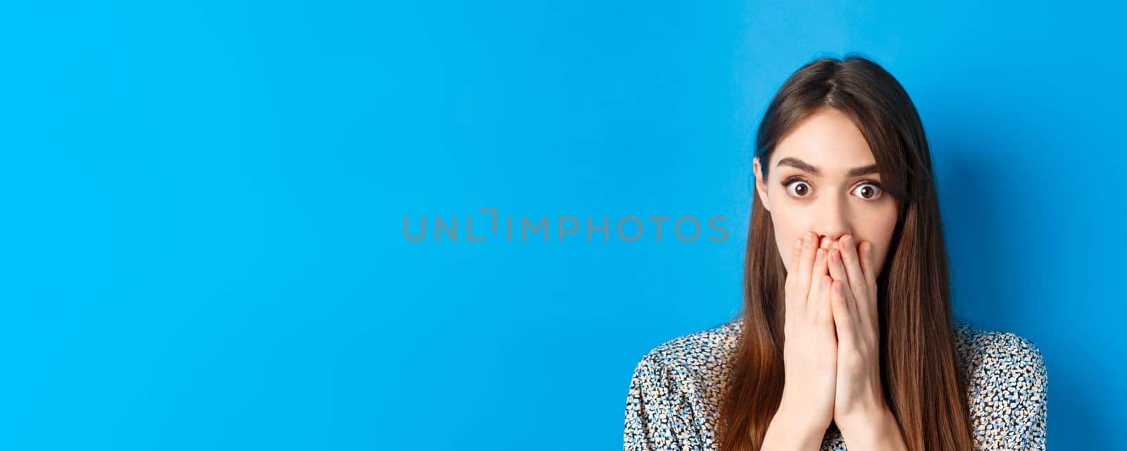 People emotions concept. Shocked young woman face, covering mouth with hands and gasping, look startled, standing on blue background by Benzoix