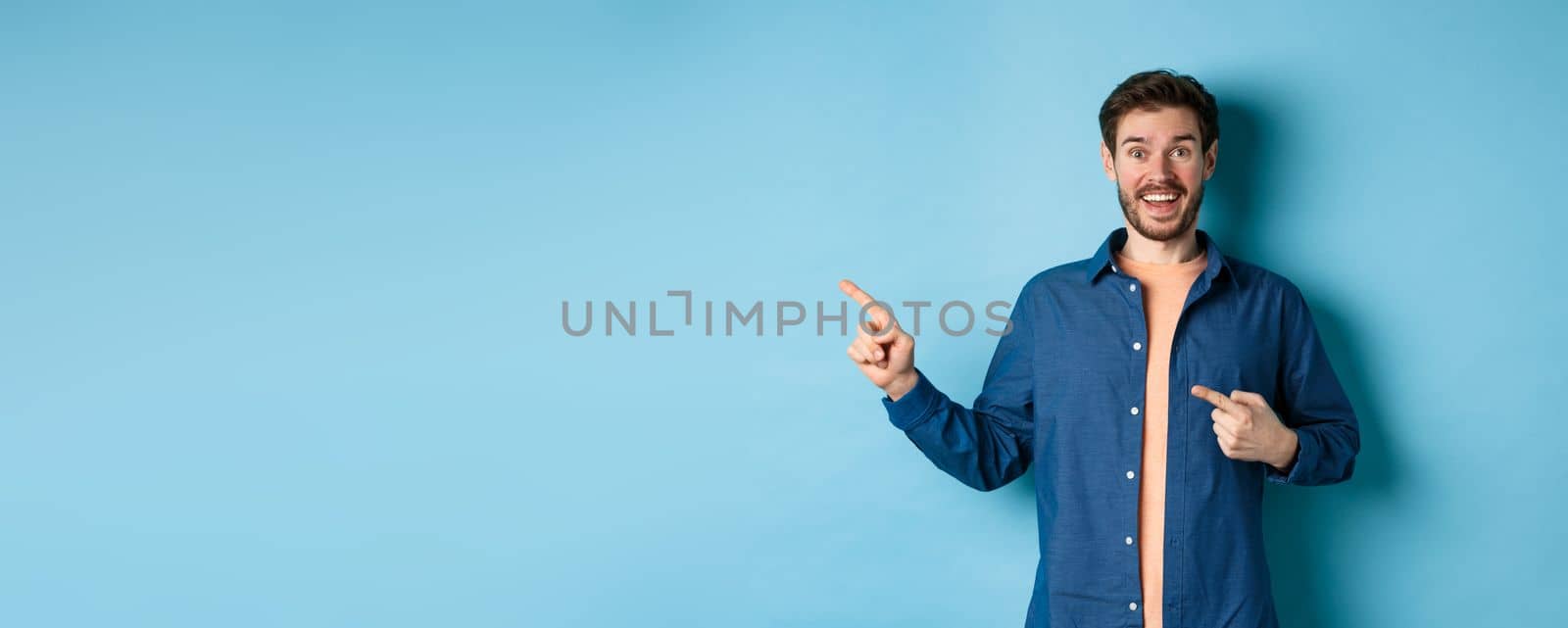 Excited and happy caucasian man smiling at camera, pointing fingers left at empty space, showing logo, standing on blue background. Copy space