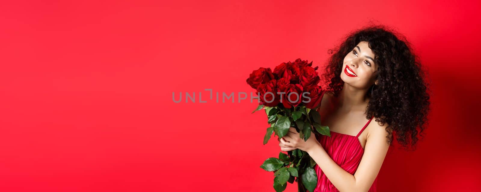 Happy beautiful woman in dress, holding flowers and smiling romantic, looking aside at logo, standing against red background by Benzoix