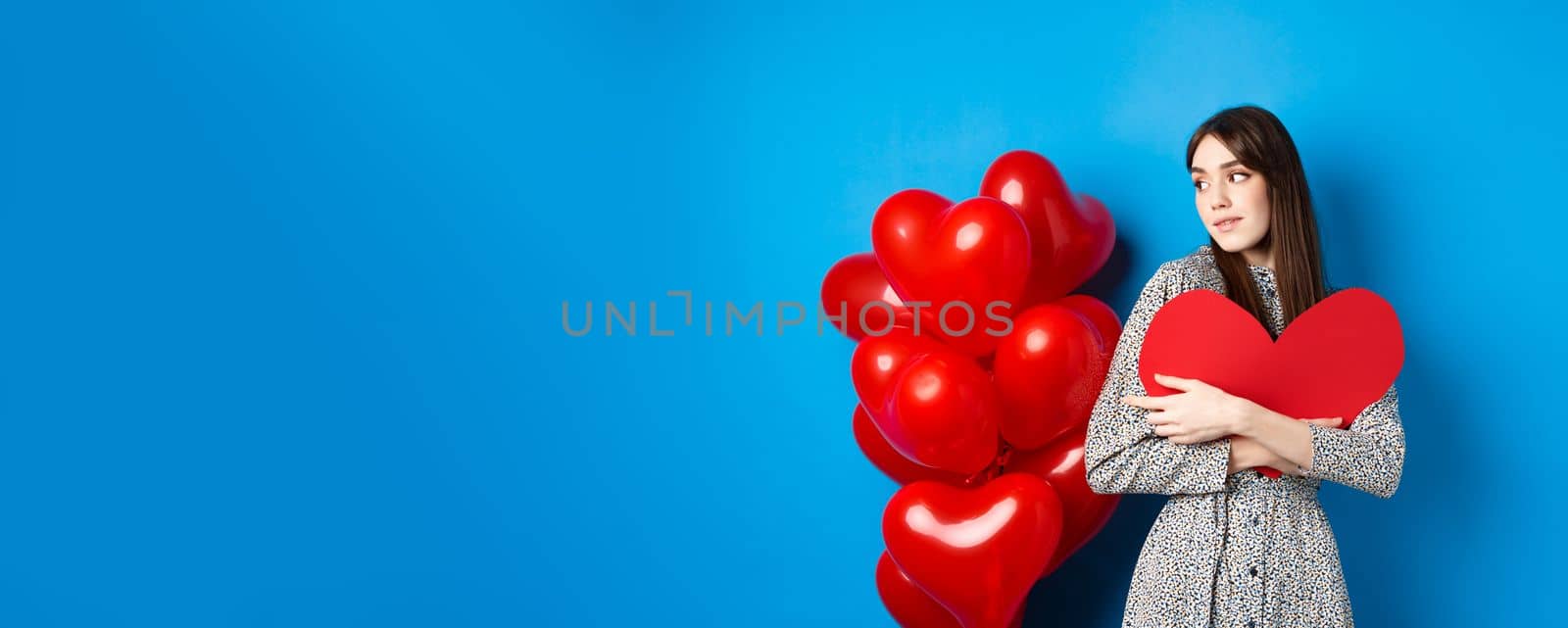 Valentines day. Beautiful and romantic woman looking pensive at balloons, hugging big red heart and smiling, waiting for love, blue background.
