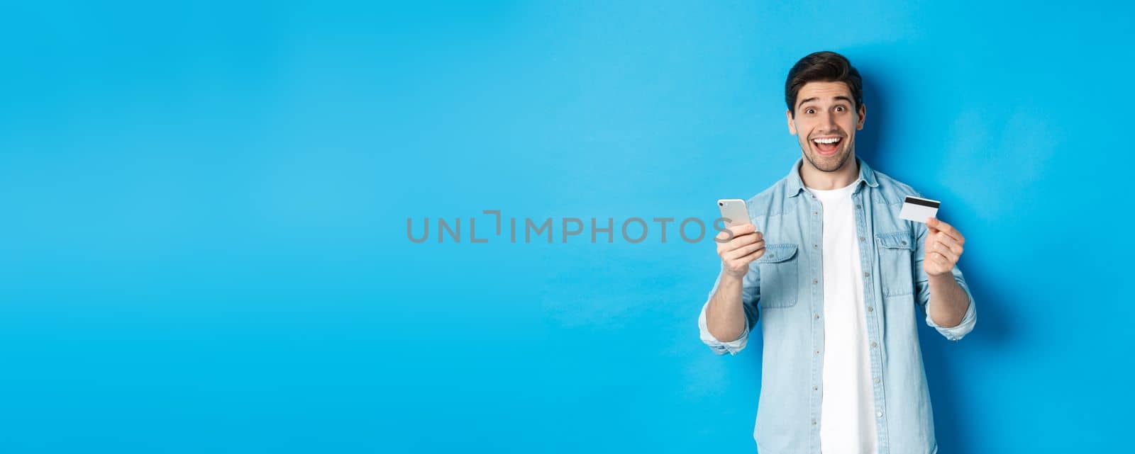 Amazed handsome man shopping online, holding mobile phone and credit card, smiling while paying for internet purchase, standing over blue background.